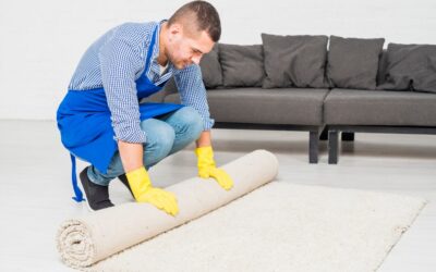 Man in blue apron and yellow gloves rolling up a carpet in a living room setting, emphasizing home cleaning and carpet maintenance.