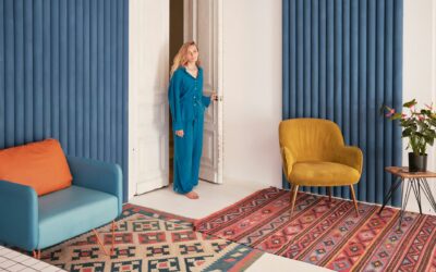 Woman in blue outfit standing at door in modern living room with colorful area rugs, blue and orange furniture, and potted plant.