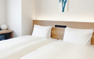 Modern hotel room with two neatly made twin beds, light-colored bedding, and a wooden headboard, featuring a bedside table and a decorative wall art piece.
